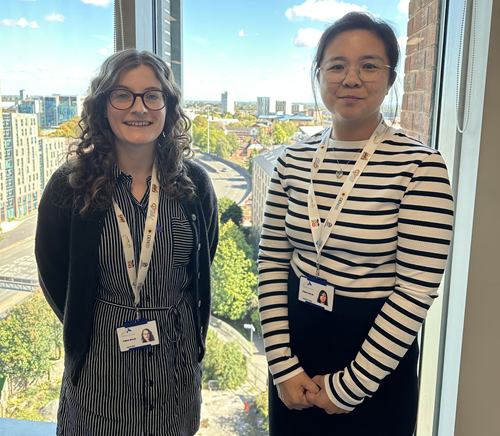 Photograph of Lauren, a woman in her thirties with long, wavy brown hair, and her colleague Seeman. They are both wearing black-and-white striped clothing and round glasses. They are both smiling directly at the camera.