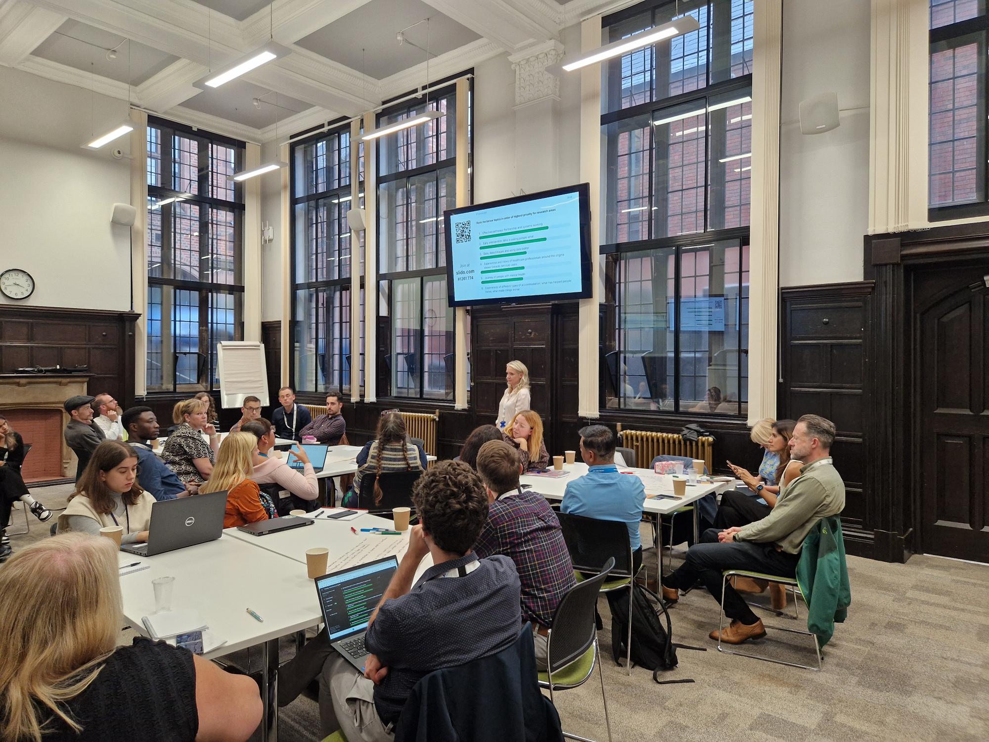 A photograph of a workshop in progress. 22 people are sitting around tables looking at a presenter standing in front of them.