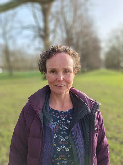A photo of Corinne Muir, standing outside with a blurry nature background, smiling at the camera