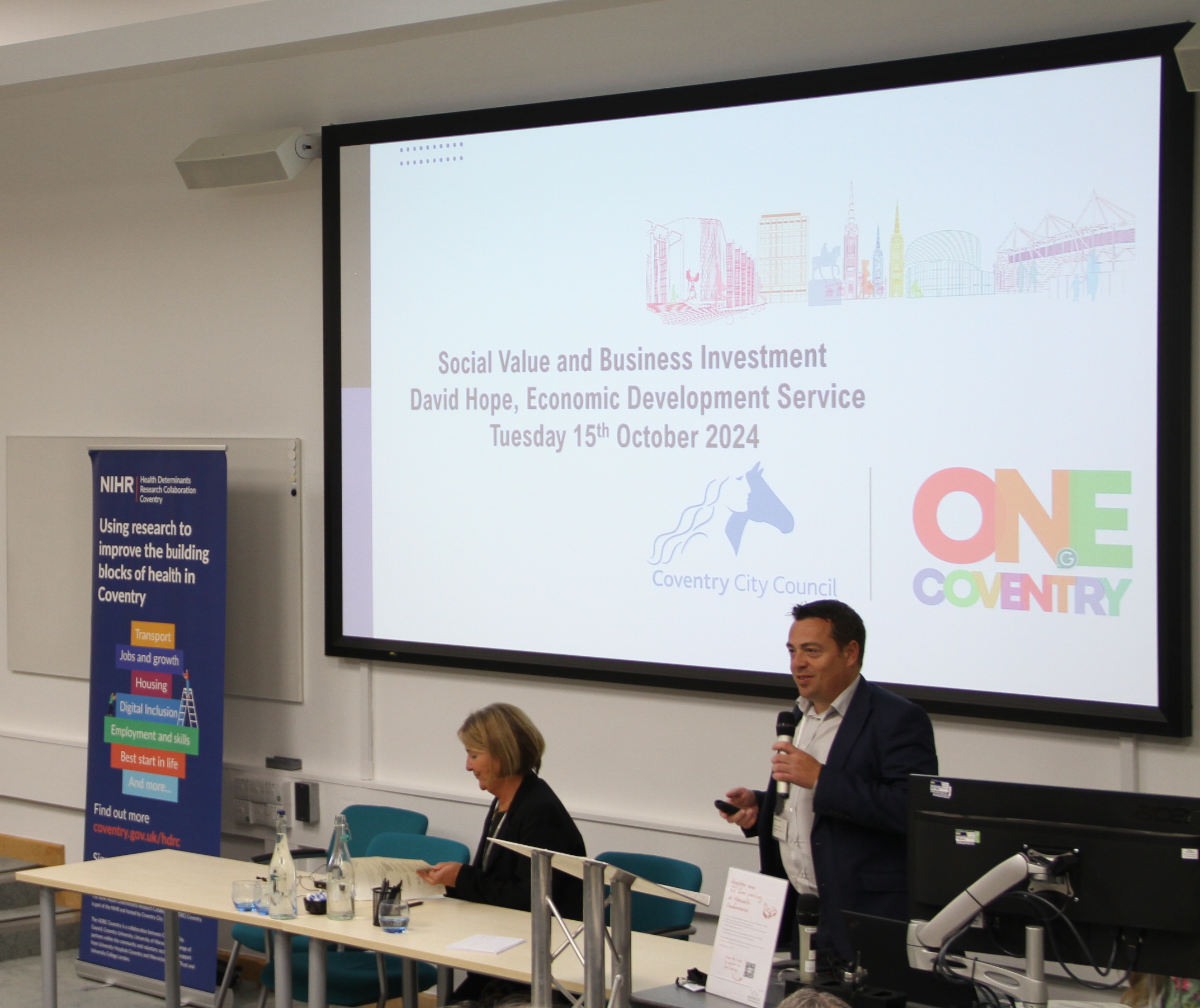 A photograph of David Hope, HDRC Research Ambassador, who is giving a talk in one of the University of Warwick's auditoriums. In front of him is a screen displaying the title 'Social Value and Business Investment'.