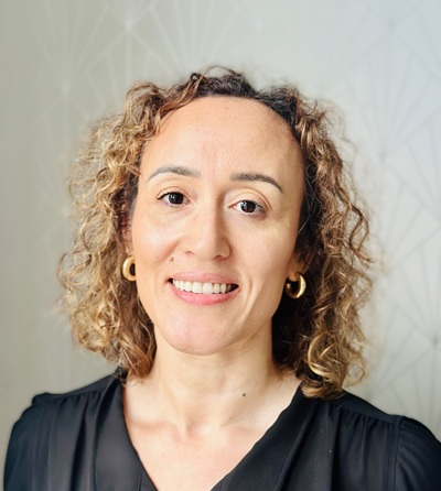 A pohoto of Michelle Simpson, wearing a black blouse and round golden earrings, smiling at the camera in front of a white background