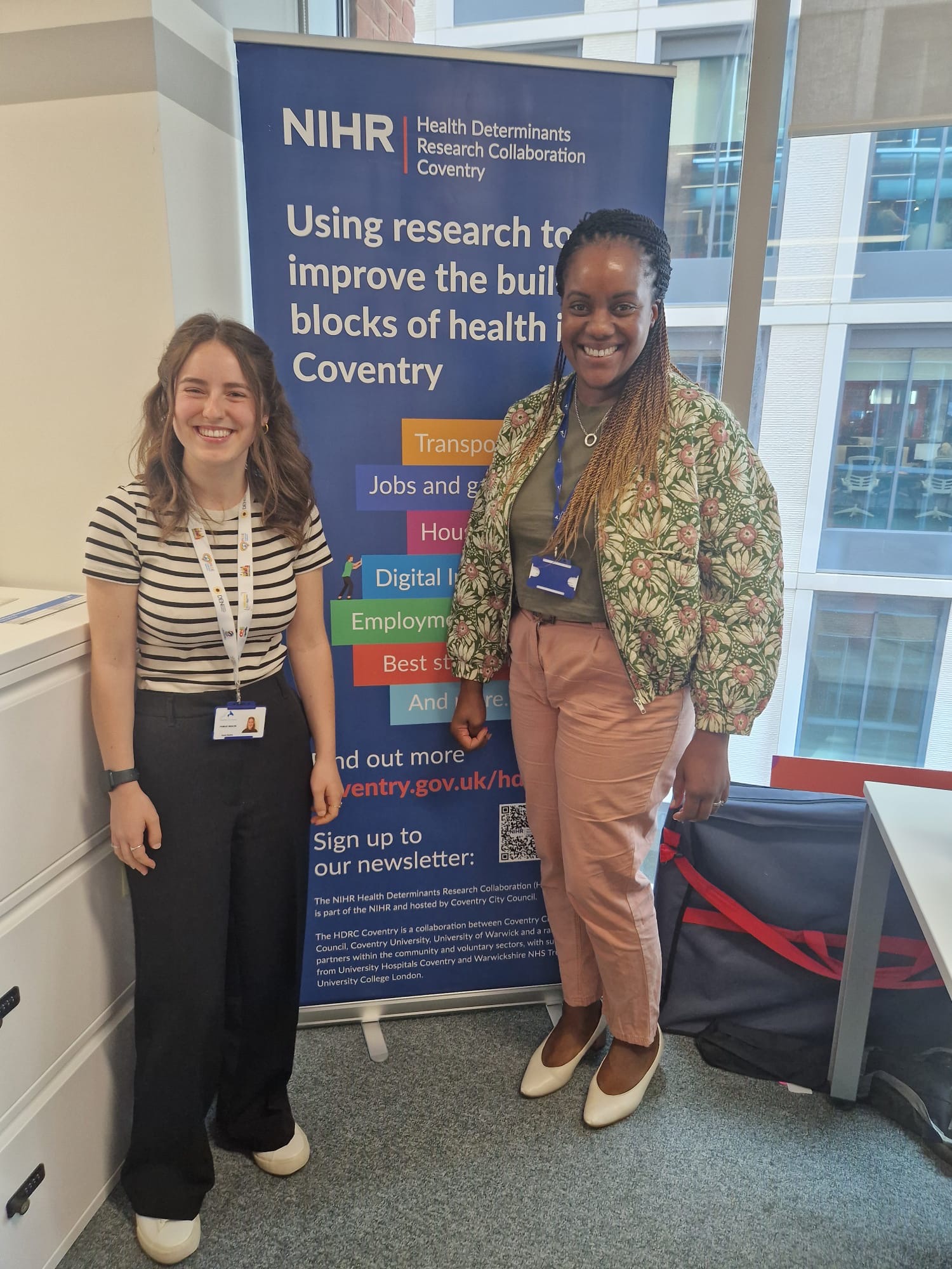 Photograph of Jessica Weeks and Kezia Davies. Kezia is a white woman in her twenties with medium-length brown hair. Jessica and Kezia are standing in front of an NIHR HDRC banner and smiling.